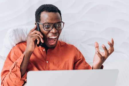 Excited African-American man talks on phone at laptop in bedの写真素材