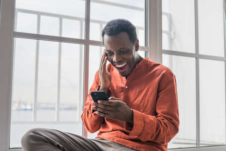 Surprised African-American man looks at smartphone by windowの写真素材