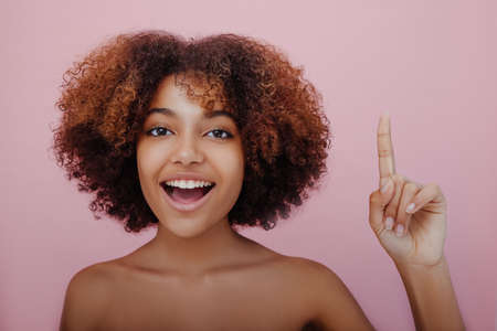 Young beautiful dark skinned woman points her finger up looks at the camera and smiles, a new idea has come, inspirationの写真素材