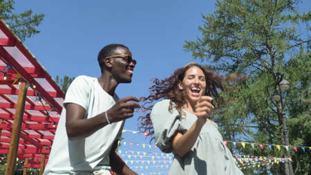 Black smiling man is dancing merrily with his girlfriend in the park on a sunny summer day against the background of colorful flagsの写真素材