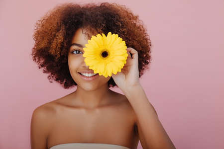 Portrait of a young beautiful African-American woman closes one eye with a yellow flower, looks at the camera and smilesの写真素材