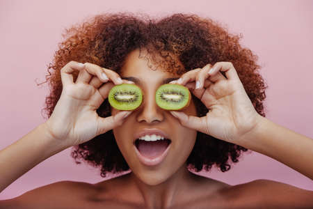 Mixed race young woman holds halves of kiwi before eyesの写真素材