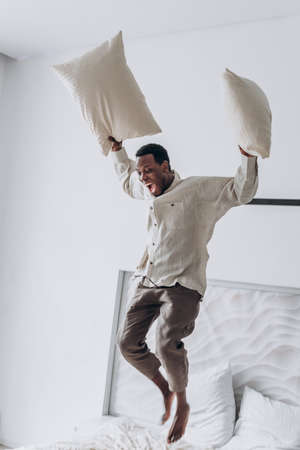 Joyful African-American man jumps throwing pillows on bedの写真素材