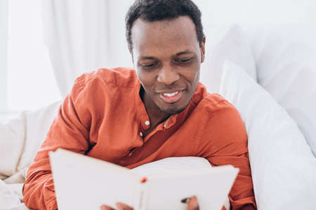 Black man in pajamas reads book lying in comfortable bedの写真素材
