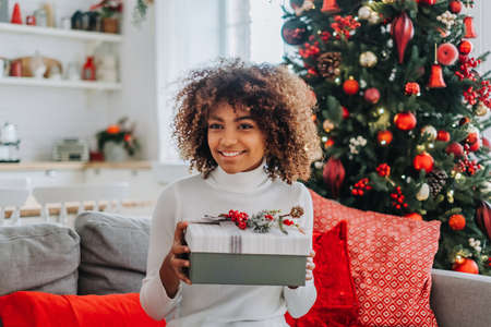 A beautiful young African American girl opens a Christmas gift at home on the sofa against the background of a festive Christmas tree.の写真素材