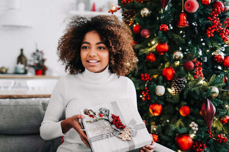 A beautiful young African American girl opens a Christmas gift at home on the sofa against the background of a festive Christmas tree.の写真素材