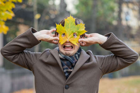Portrait of a crazy funny man in a coat covering his face with a maple leaf mask and making a funny faceの写真素材