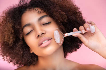 Portrait of a young well-groomed dark-skinned woman with lush hair, doing facial massage with the help of a roller with closed eyes, care and skin careの写真素材