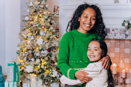 Portrait of a mother with dark curly hair and a little daughter against the background of a christmas tree, gifts and candlesの写真素材