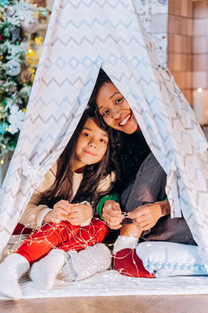 Mom and little daughter look out of the tent in the room and hold a garland in their hands against the background of a Christmas treeの写真素材