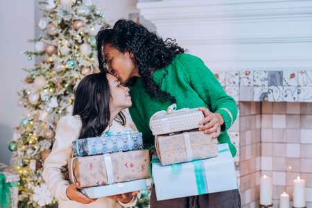 African american mom and daughter stand with christmas gifts near the christmas tree, mother kisses...