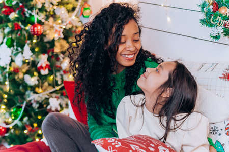 Cute mom and daughter look at each other while sitting on the bed on the background of christmas decorationsの写真素材