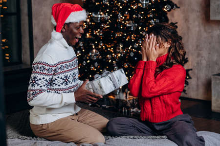 African american man in red cap gives christmas gift box surprise to mixed race woman in red sweater against new year tree background, couple celebrating at homeの写真素材