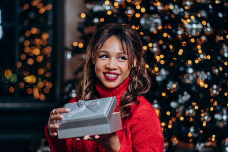 Portrait smiling mixed race woman in red sweater holding an open gift on background of a christmas...