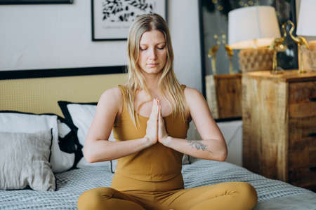 Blonde woman meditates with closed eyes sitting on bedの写真素材
