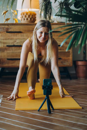 Woman with blonde hair exercises on mat with phone on tripodの写真素材