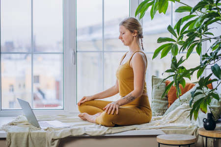 Blonde-haired woman meditates on windowsill using laptopの写真素材