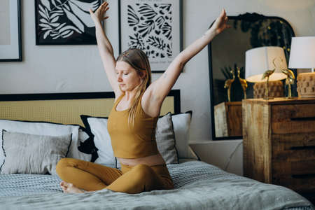 Young woman stretches sitting on bed covered with blanketの写真素材