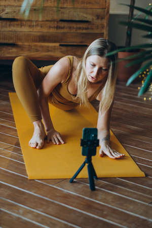 Athletic woman stretches on yellow mat with phone on tripodの写真素材