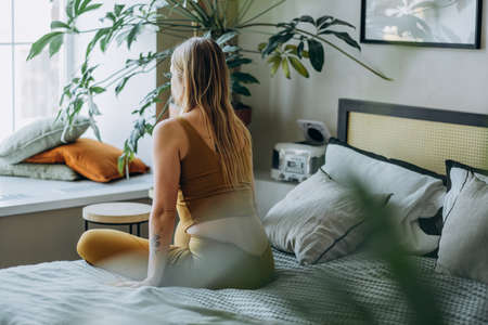 Woman with blonde hair stretches sitting on bed and looking at windowの写真素材