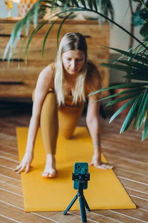 Woman with blonde hair exercises on mat with phone on tripodの写真素材