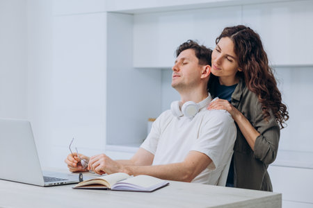 Wife massaging shoulders of tired husband freelancer working on laptop at home in kitchenの写真素材
