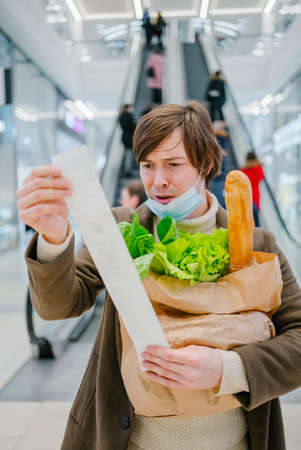 Man in a medical mask looks in horror at a check in a supermarket and holds a bag of groceriesの写真素材