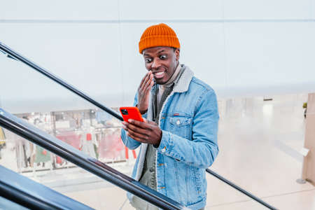 Happy African-American guy looks at phone going up escalatorの写真素材