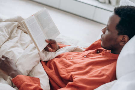 Black man in pajamas reads book lying in comfortable bedの写真素材