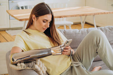 A happy woman with a bionic prosthetic arm sits on the couch with a phone and communicates online with friends or surfs the Internet or makes online purchasesの写真素材