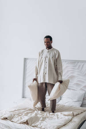 Joyful African-American man jumps throwing pillows on bedの写真素材