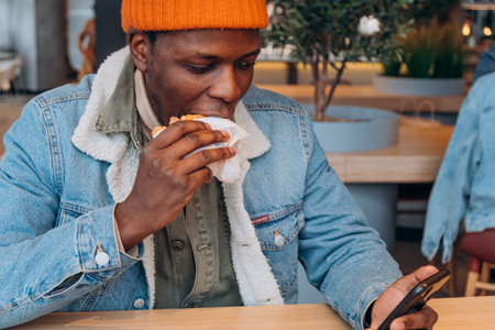 African-American guy eats tasty hamburger looking into phoneの写真素材