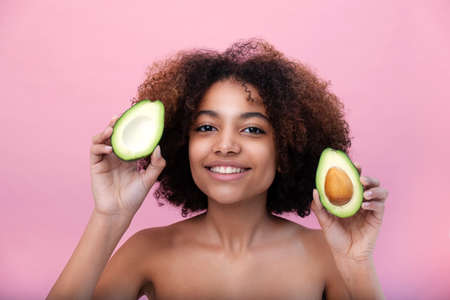 Portrait of a young beautiful African American woman with lush hair holding a sliced avocado on a pink backgroundの写真素材