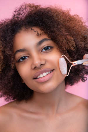 Portrait of a young well-groomed dark-skinned woman with lush hair, doing facial massage with the help of a roller with closed eyes, care and skin careの写真素材