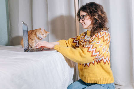Stylish female freelancer working at home on a laptop sitting on floor with her pet red cat.の写真素材