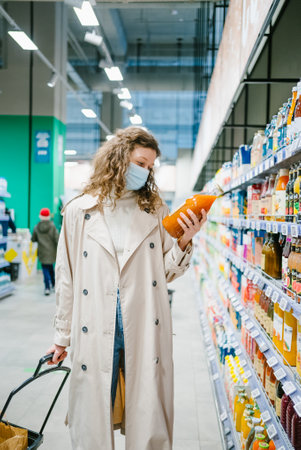 A young girl in a raincoat in a medical mask reads the composition on a glass bottle of juice in a supermarketの写真素材