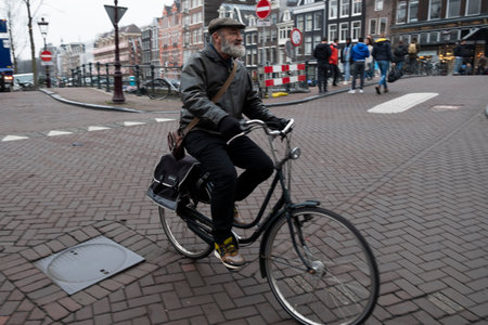 An elderly man with a beard, wearing a hat and a jacket, rides a bicycle across a cobblestone street in an urban European setting.のeditorial素材