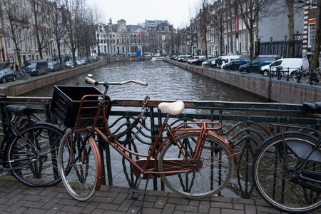 Bicycles parked along a canal railing in Amsterdam, with historic buildings lining the waterway under a grey sky.のeditorial素材
