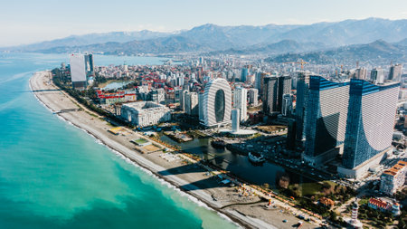 Aerial shot captures a modern cityscape along the coast, featuring striking skyscrapers juxtaposed with the serene blue sea and backed by majestic mountains.のeditorial素材