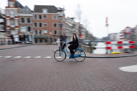 Motion blur captures the swift movement of a cyclist on a bright blue bike, with Amsterdams canal and streetscape in the background.のeditorial素材