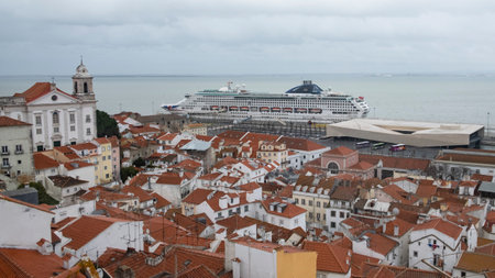 Overlooking the terracotta rooftops of Lisbons historic district, a large cruise ship is docked by the Tagus River, under a cloudy sky.のeditorial素材
