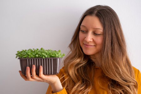 A contented young woman in a mustard yellow top admires a pot of lush green microgreens shes holding gently in her hands, embodying wholesome living.の写真素材
