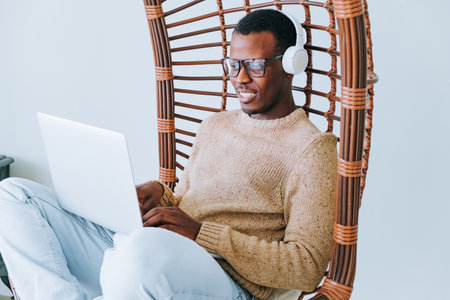 Young African American Man Working on Laptop While Sitting in a Wicker Chair With Headphonesの写真素材