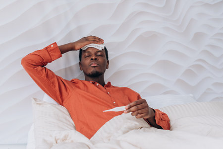 African American Man with fever resting in bed, checking temperature with thermometer, holding wet cloth to forehead, wearing orange shirtの写真素材