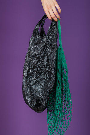 Woman Hand Holding Black Plastic Bag and Green Mesh Bag Against Purple Background