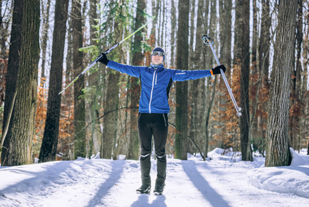 Joyful cross-country skier in blue jacket stands with arms raised, holding skis and poles in snowy...