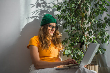 Woman in green beanie and yellow shirt works on her laptop in sunny room next to leafy plant.