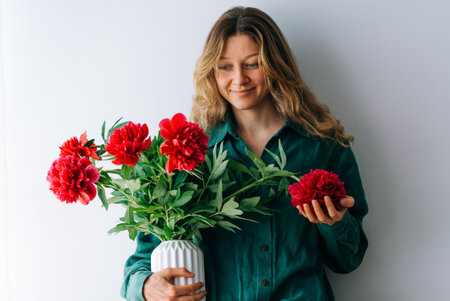 Smiling woman in green shirt holding vase with red flowers pions, against white background.の写真素材