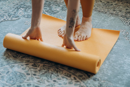 Close-up of a woman unrolling a yellow yoga mat on a patterned rug, preparing for a yoga session at...