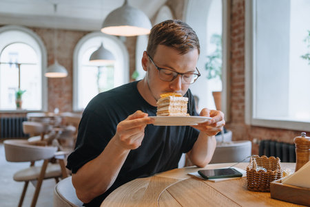 Man with glasses sniffs delicious layered piece of cake while sitting at table in cozy cafe.